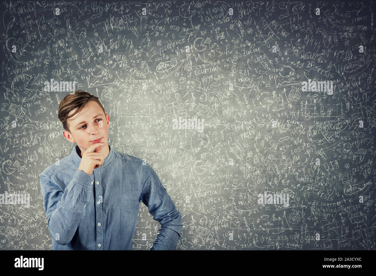 Smart teenage boy thinking, keeps hand under chin, stands in front of a blackboard solving hard mathematics calculation and equations. Puzzled student Stock Photo