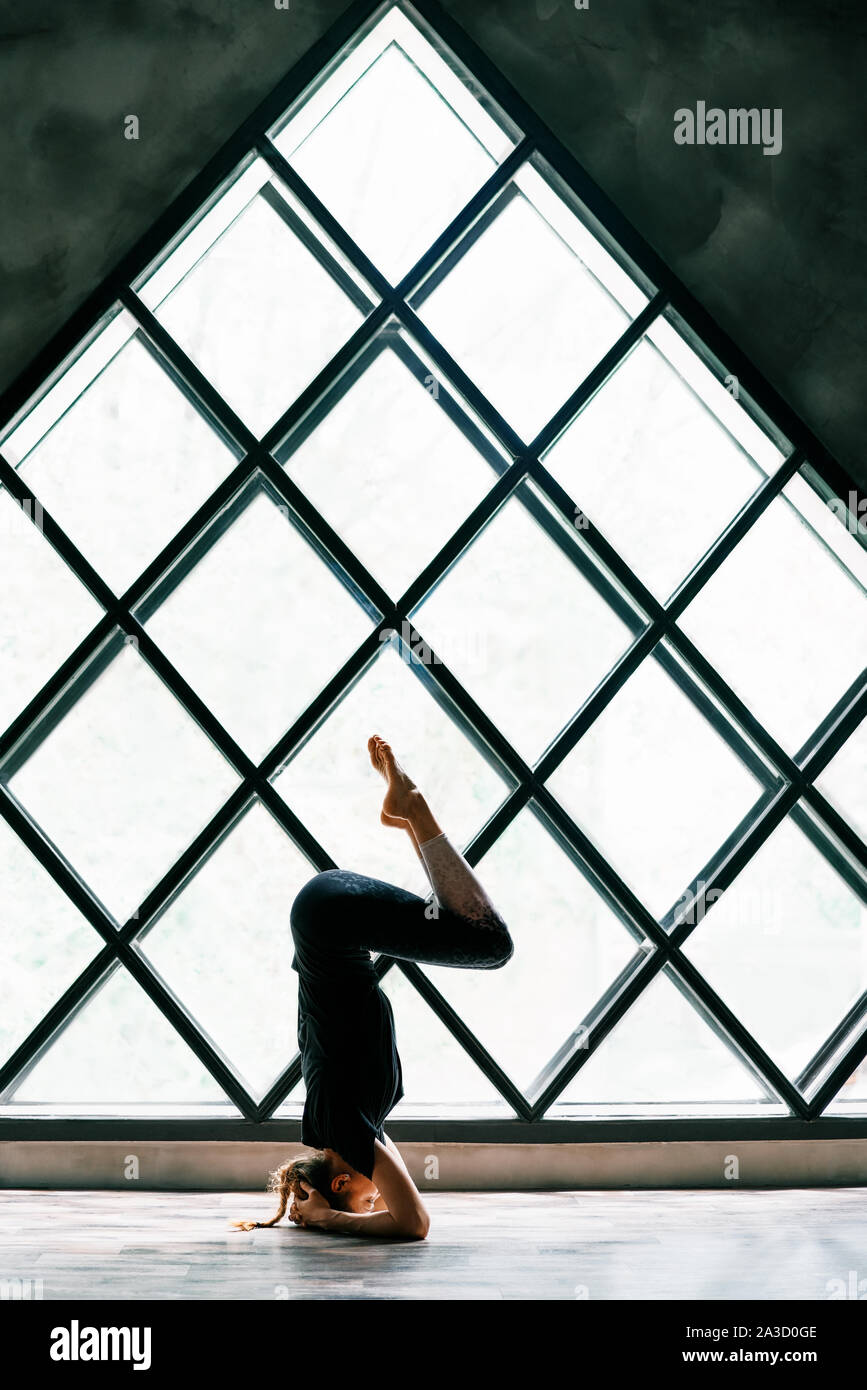 Young attractive woman practicing yoga standing in supported headstand pose, akunchanasana, on large triangular window background. Healthy lifestyle c Stock Photo