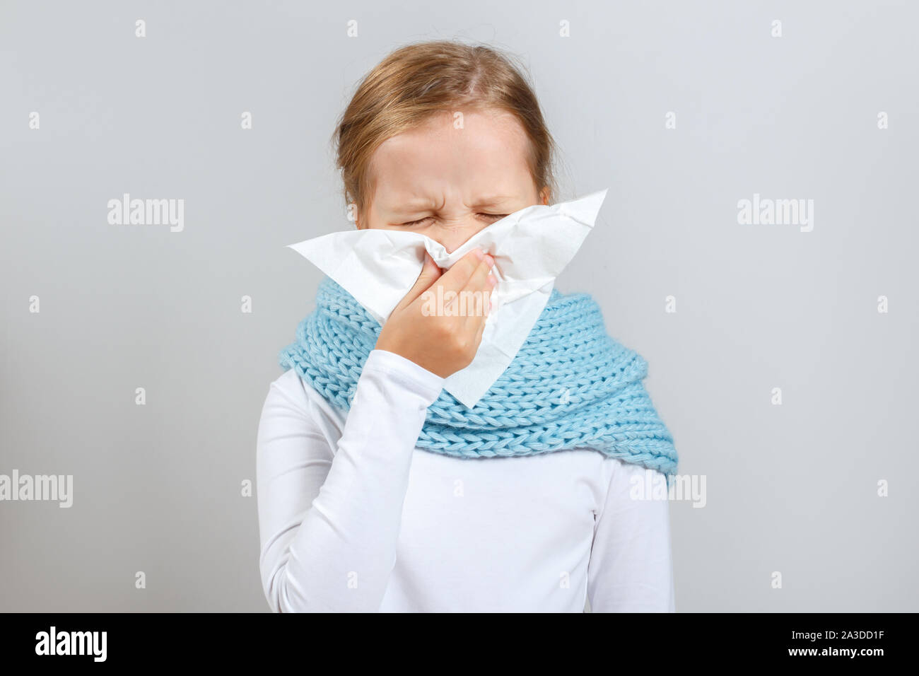 Cold season. Little girl in a warm scarf blows her nose. A child on a gray background. Stock Photo