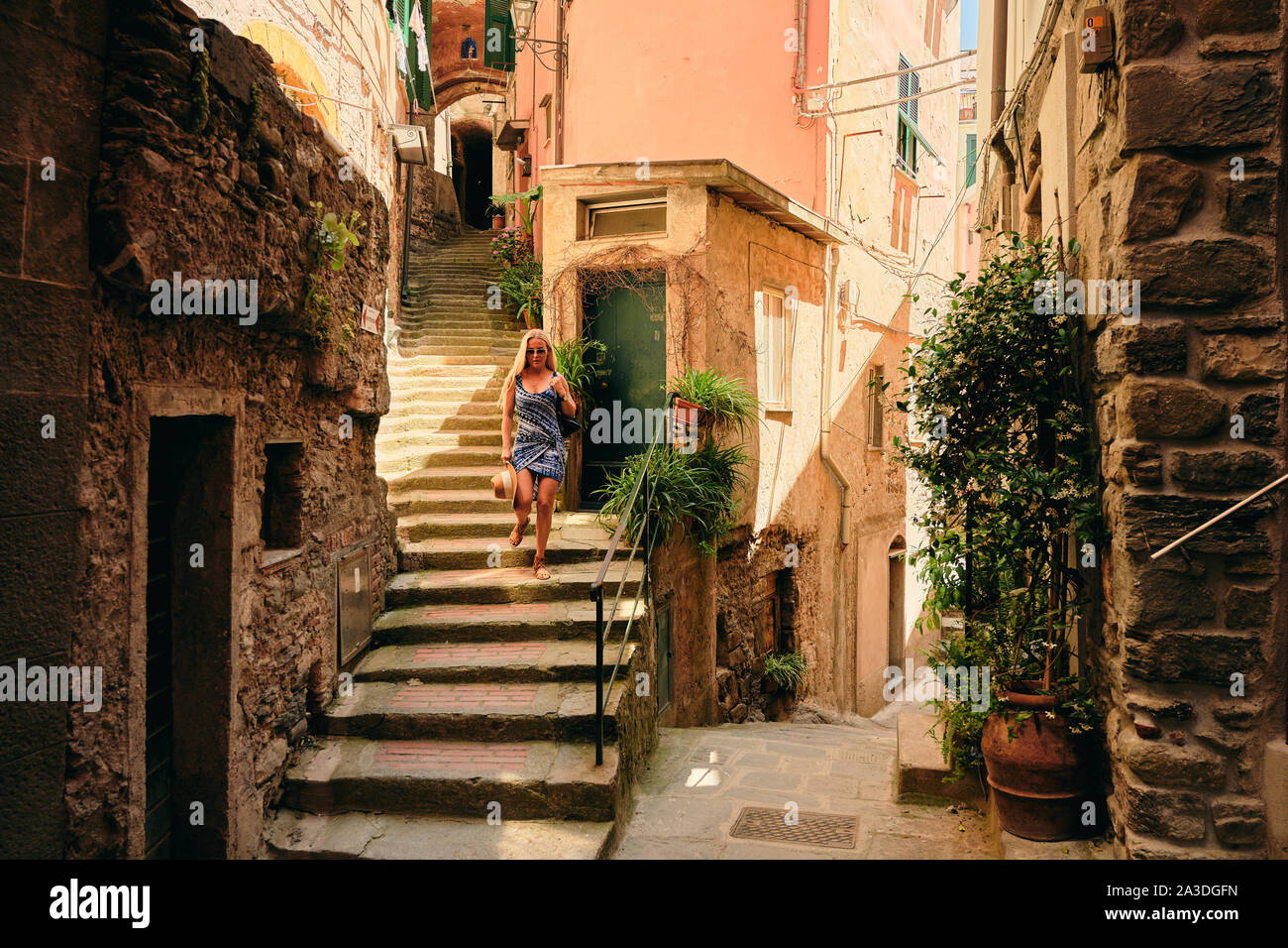 Relaxed woman in sundress and sunglasses walking along staircase down narrow sunny street at rural town Stock Photo