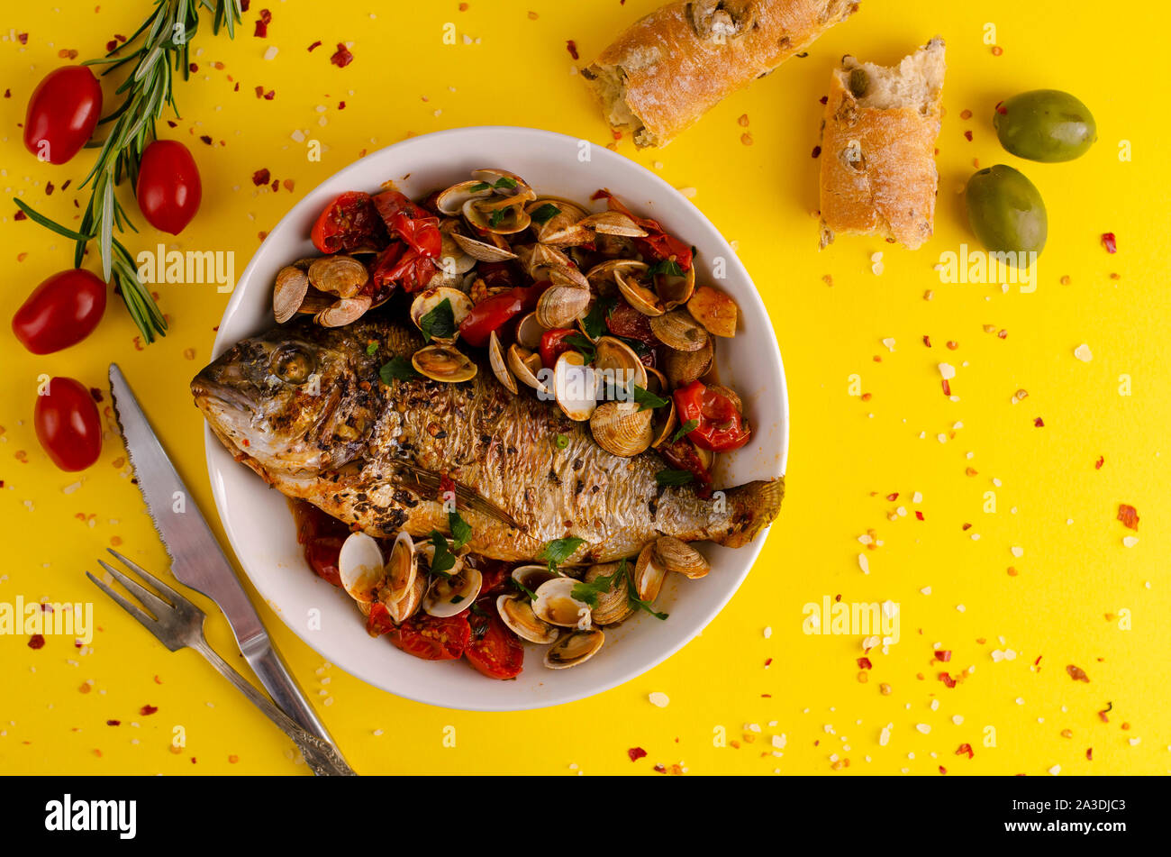 Cooked Sea bream with clams in a white bowl, olive bread and tomatoes on yellow background. Seafood concept, overhead shot,copy space Stock Photo