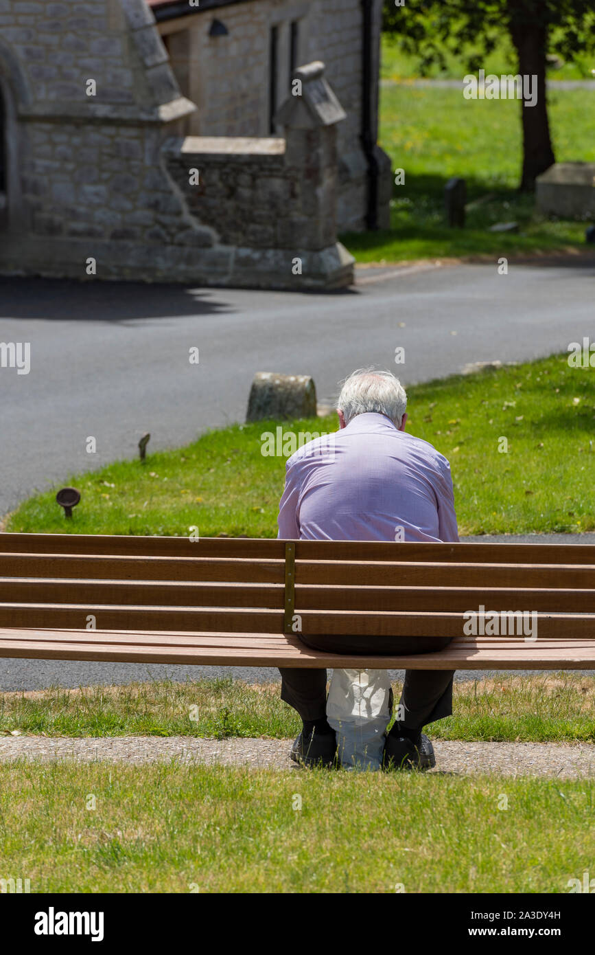 An elderly grey haired man or pensioner sitting on a wooden bench in a cemetery or graveyard near a church lonely and on his own. Stock Photo