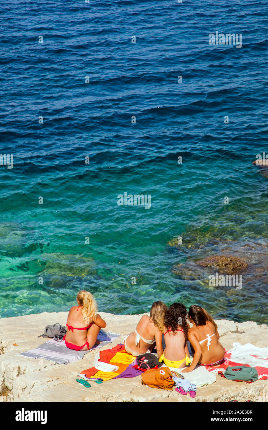 Young women sitting talking and chatting while sunbathing in bikinis on rocks at the beach at  Greek island holiday resort  of kassiopi Corfu Greece Stock Photo
