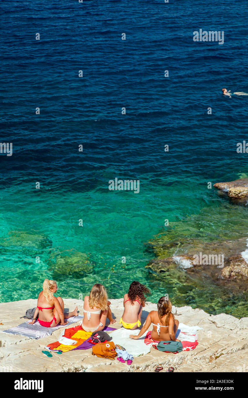 Young women sitting talking and chatting while sunbathing in bikinis on rocks at the beach at  Greek island holiday resort  of kassiopi Corfu Greece Stock Photo