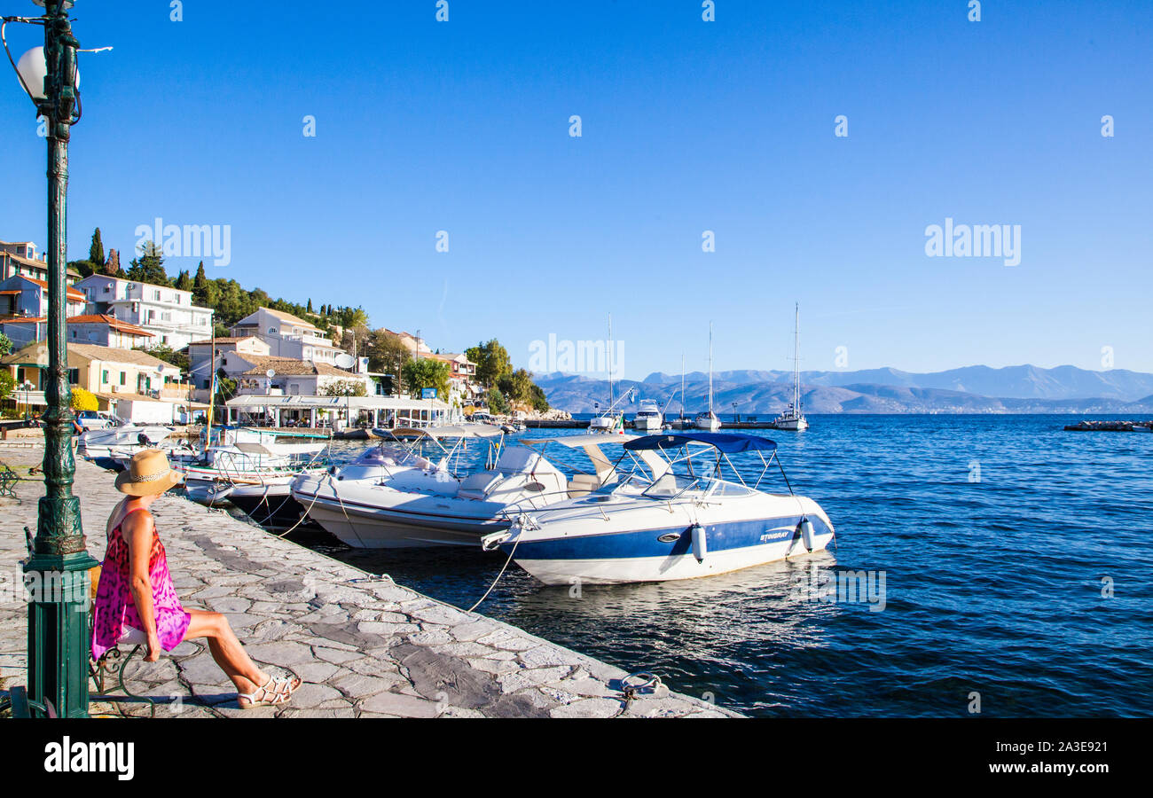 Woman tourist holidaymaker sitting by the harbour in the Greek holiday resort of Kassiopi corfu Greece with boats in sea and blue sky skies Stock Photo