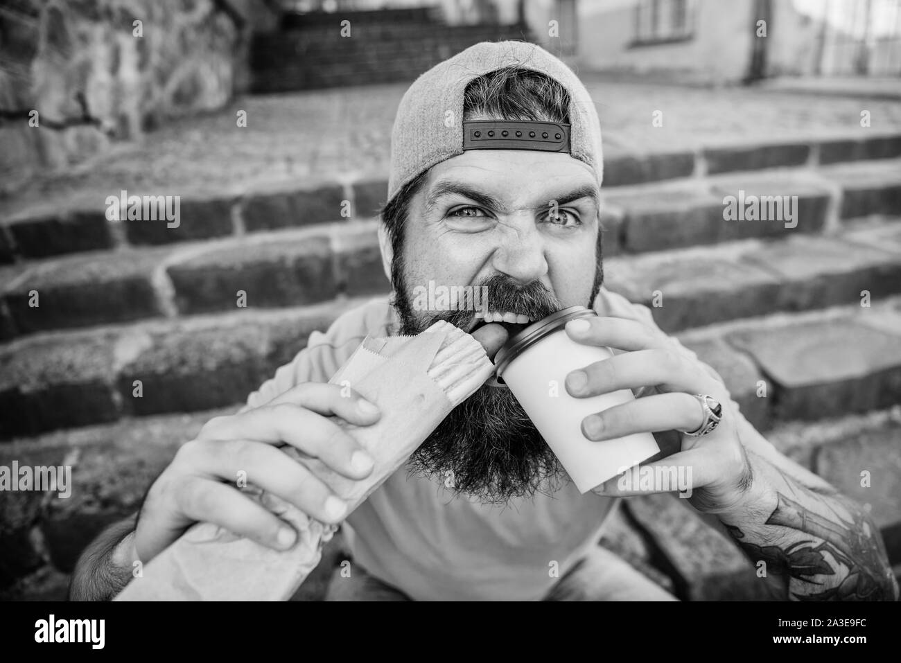 Snack for good mood. Guy eating hot dog. Street food concept. Man bearded eat tasty sausage and drink paper cup. Urban lifestyle nutrition. Junk food. Carefree hipster eat junk food while sit stairs. Stock Photo
