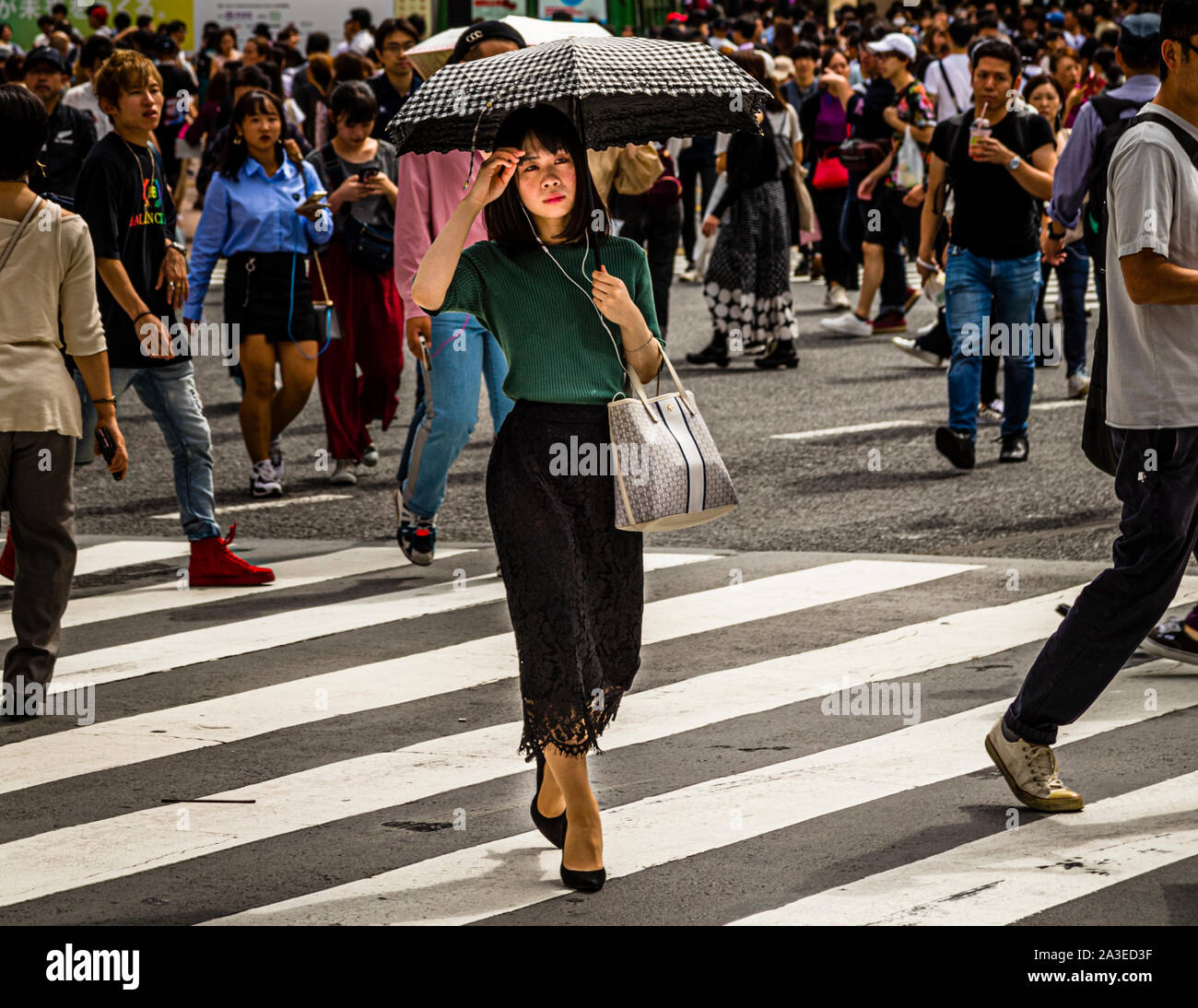 A pedestrian with a checkered parasol crosses the famous Shibuya scramble crossing in Tokyo, Japan. Numerous passers-by crowd the background. The busy crossing is considered one of the world's most frequented pedestrian crossings. Stock Photo
