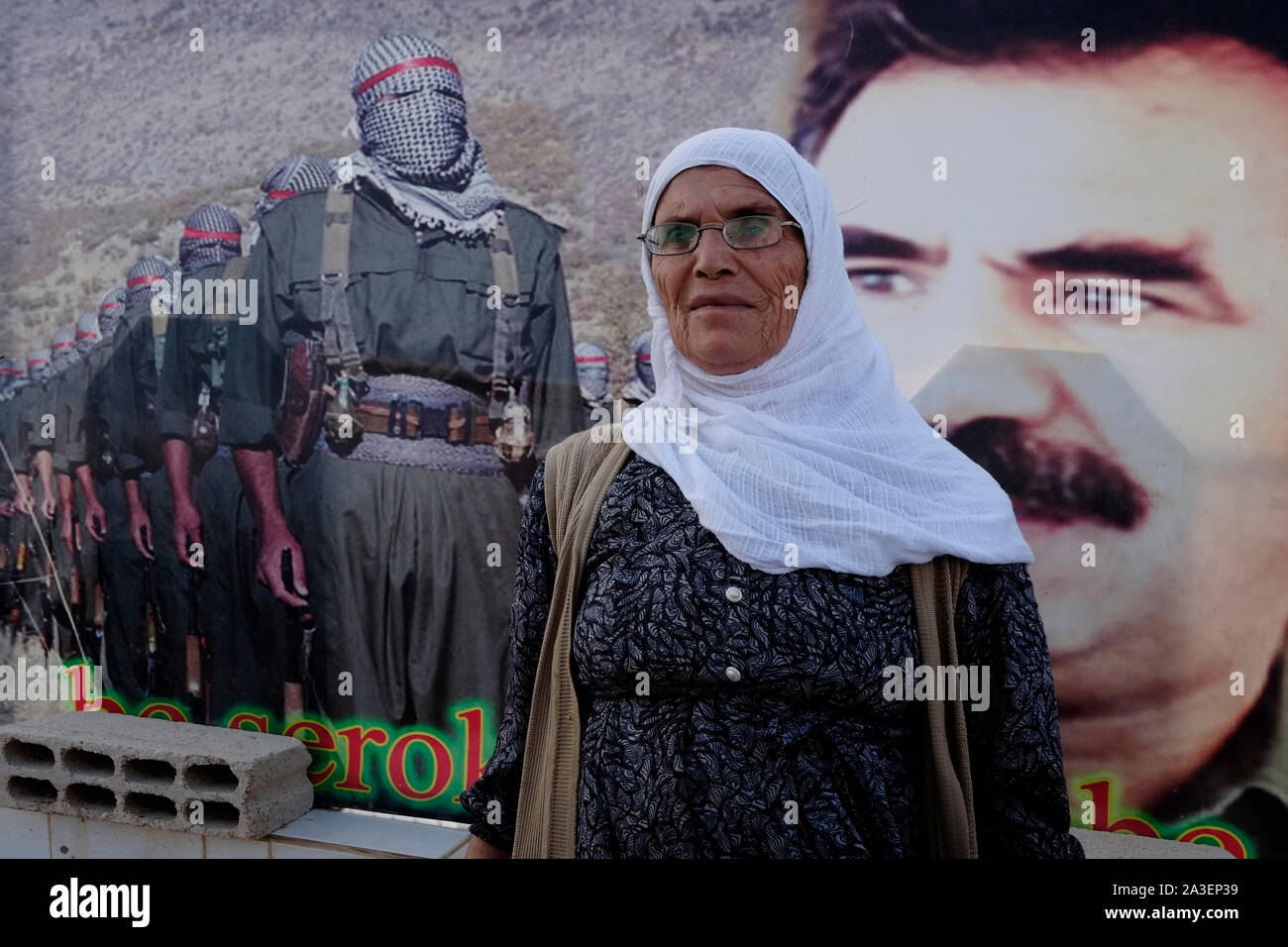 An elderly Kurdish woman stands next to a poster showing veiled YPG Kurdish fighters and the former PKK militant leader Abdullah Ocalan in Al Hasakah or Hassakeh district in Rojava the de facto Kurdish autonomous region originating in and consisting of three self-governing cantons in northern Syria Stock Photo