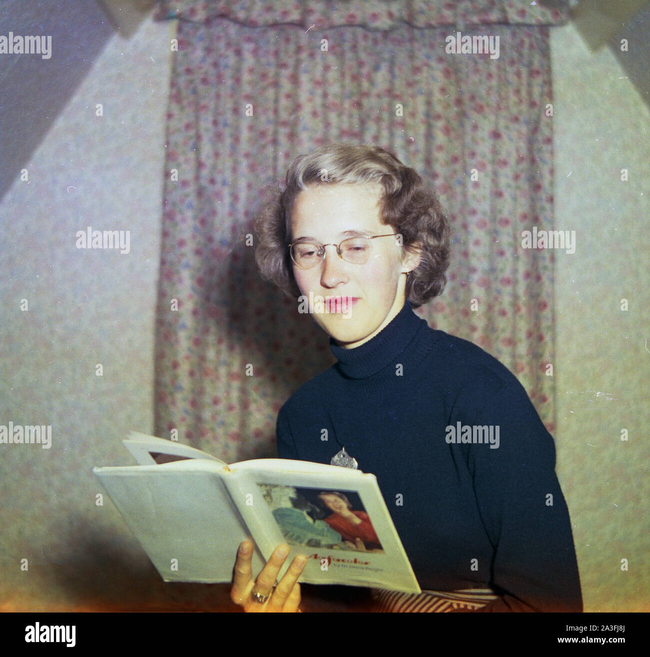 1953, historical, an Elegant looking English lady wearing a blue, woollen roll-neck top and glasses inside a room reading a reference book about Agfacolor by Agfa technician Dr. Heninz Berger. Agfacolor was the name of a series of color film products made by Agfa of Germany and the book was an english translation. Founded in Berlin in 1860s, Agfa entered the field of photograhy in 1898. Stock Photo