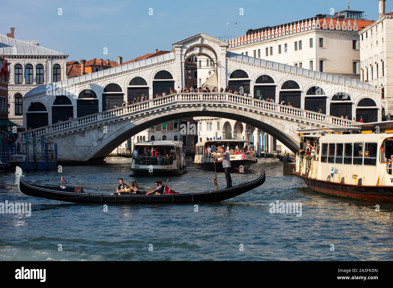 Rialto Bridge in Venice, Italy, spanning the Grand Canal with gondolas and vaporetto boats passing underneath.