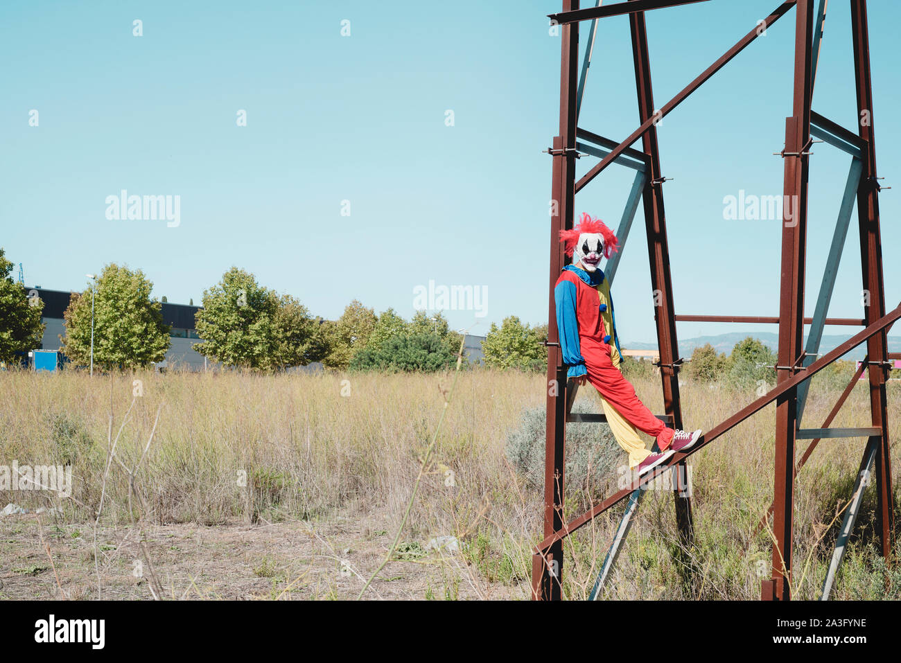 a scary clown wearing a colorful yellow, red and blue costume outdoors, hanging from the rusty structure of an abandoned billboard Stock Photo