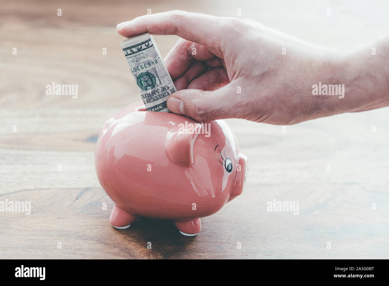 close-up of male hand putting folded dollar bill in pink piggy bank on wooden table, home finance and saving money concept Stock Photo
