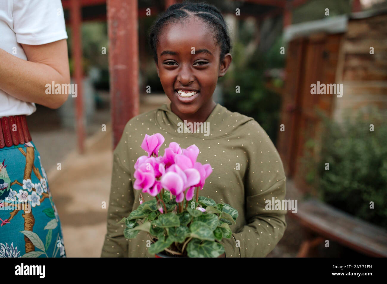 Smiling black girl holding plant with pink flowers at nursery Stock Photo