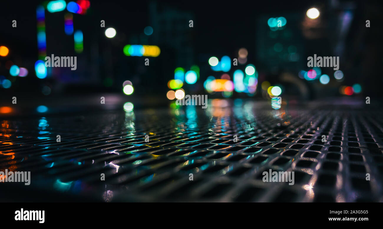 Abstract dark panoramic night city photo with shiny wet street sewer grate on urban road and blurred lights on a background, close-up photo with selec Stock Photo
