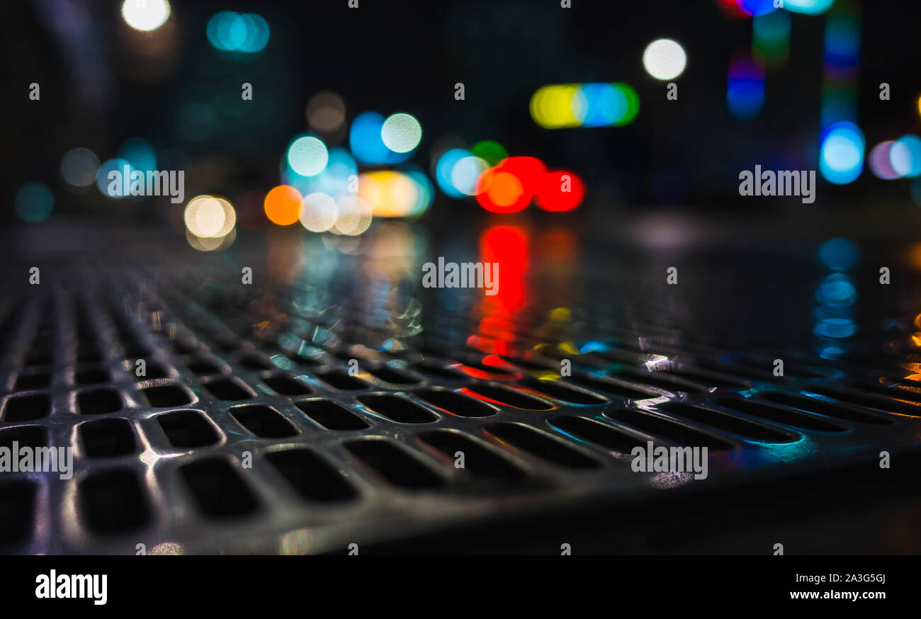 Abstract night city photo with shiny wet street sewer grate on urban road and blurred lights on a background, close-up photo with selective focus Stock Photo