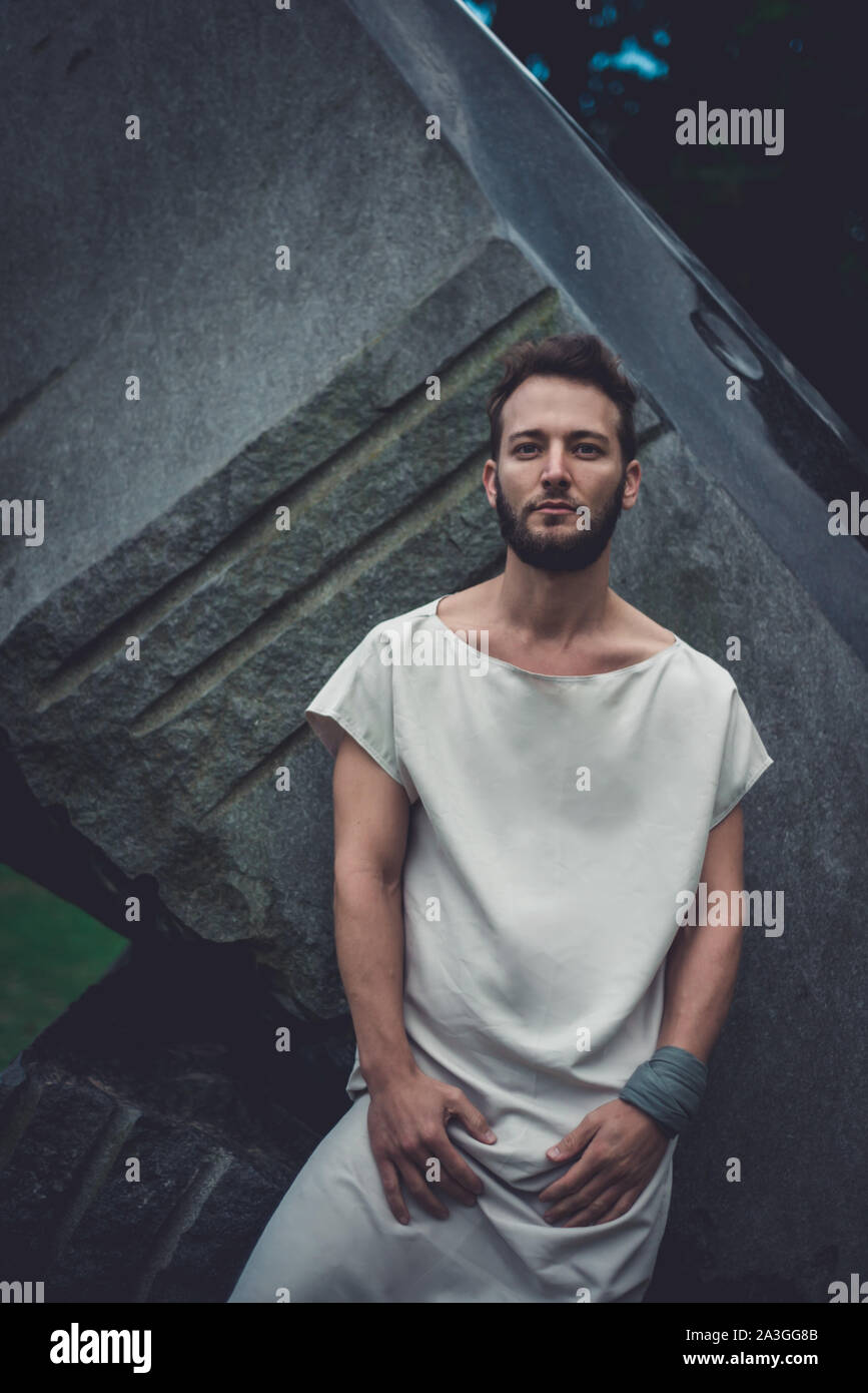 Moody portrait man in mid-thirties with an intense gaze leaning up a large dark stone structure. Stock Photo