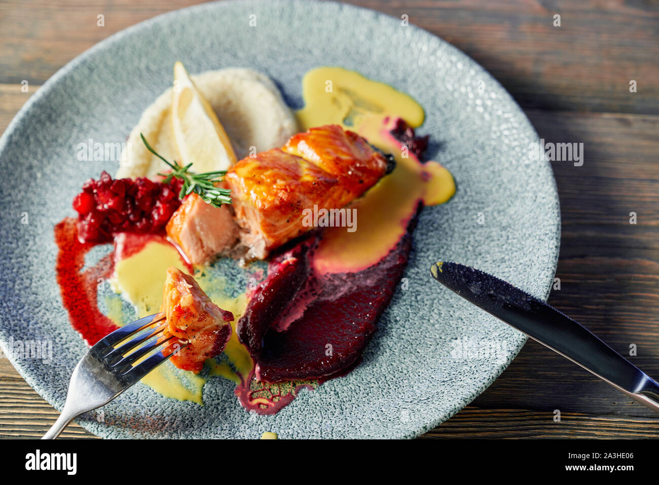 View from above of delicious grilled fish served with fruit garnish on plate. Fork with piece of meat and knife. Tasty meal with sauce and lemon. Concept of diet and vitamin. Stock Photo