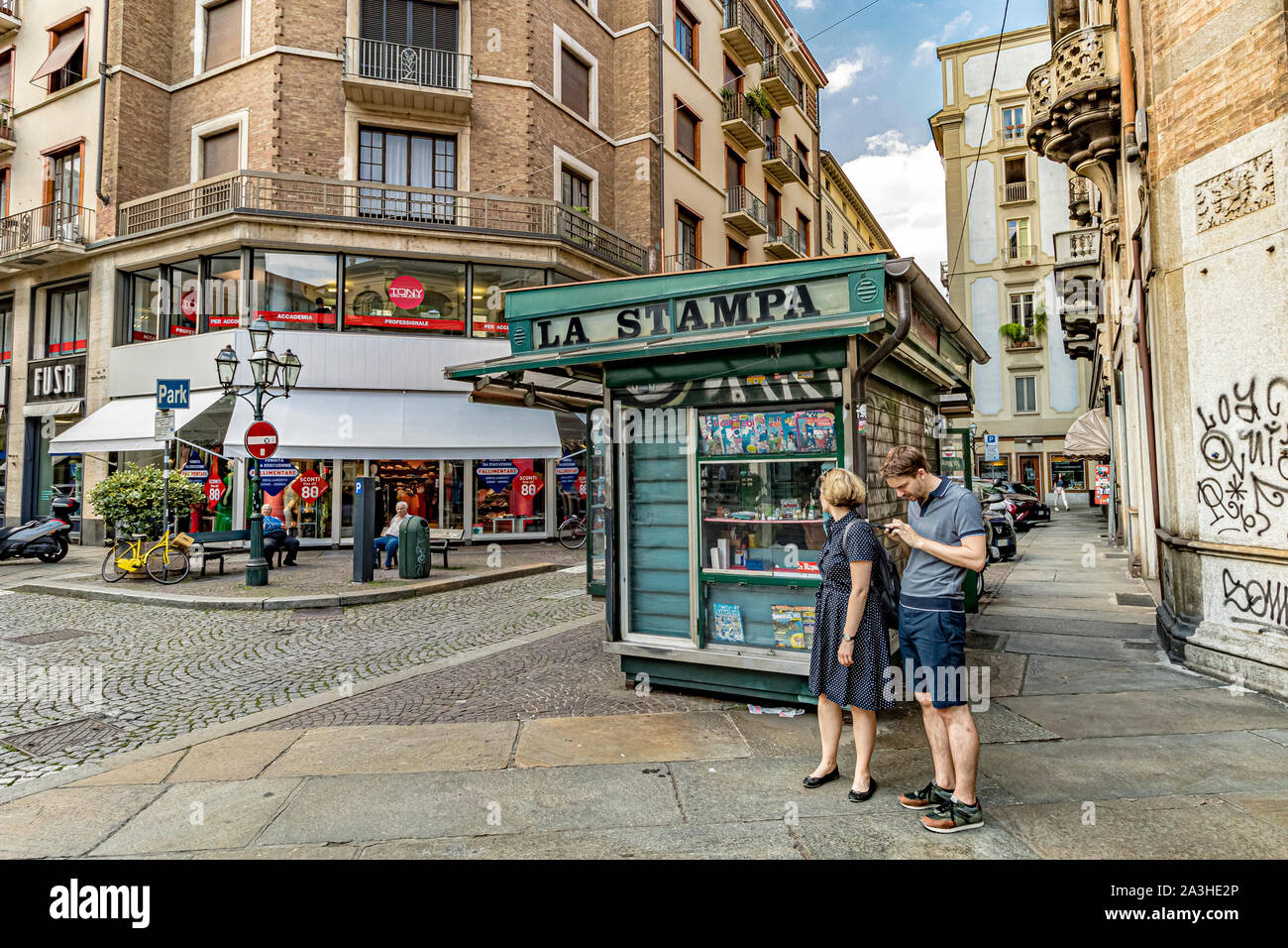 A couple standing next to a green La Stampa newspaper kiosk whilst the man looks at his mobile phone  on Via Pietro Micca ,Turin.Italy Stock Photo