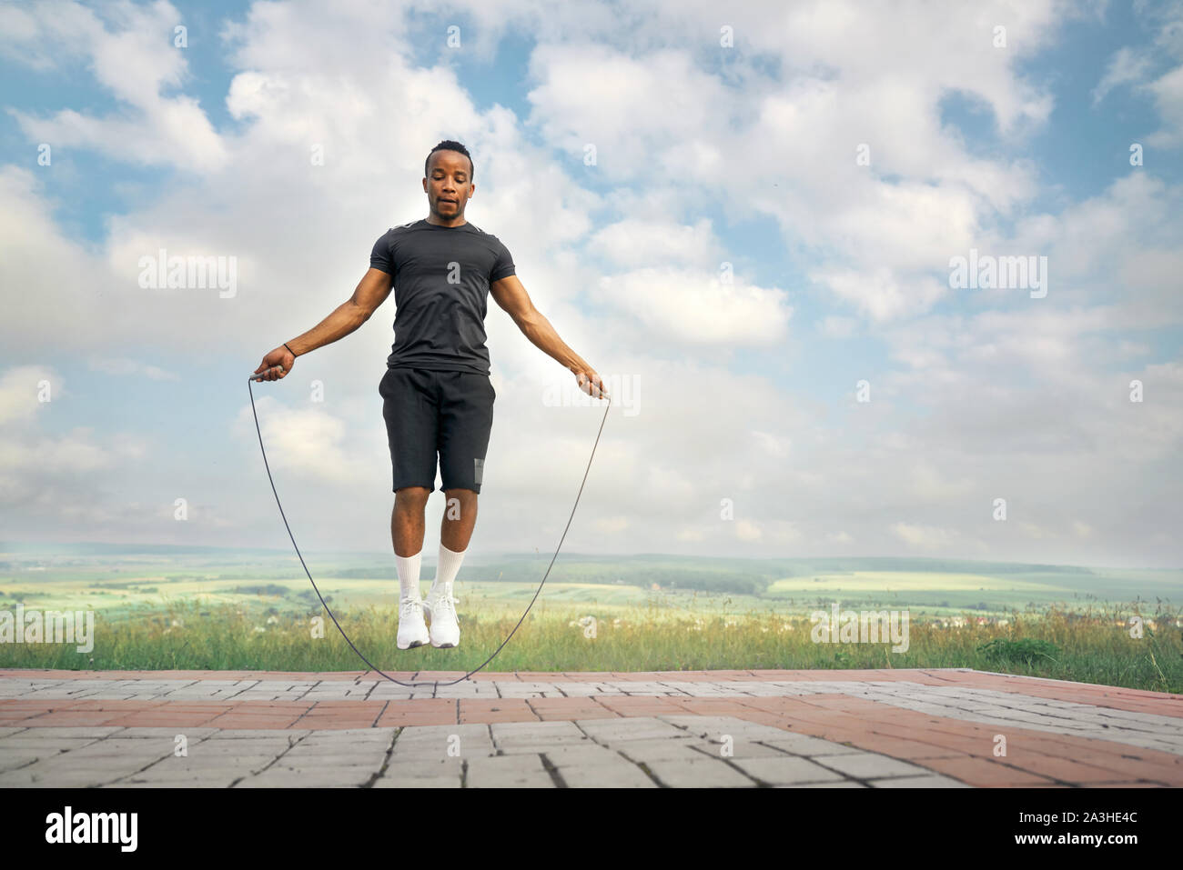 Handsome, strong sportsman in black t shirt and shorts training with jumping rope outdoors. Active athlete with sportive body doing exercises on background of blue sky. Stock Photo
