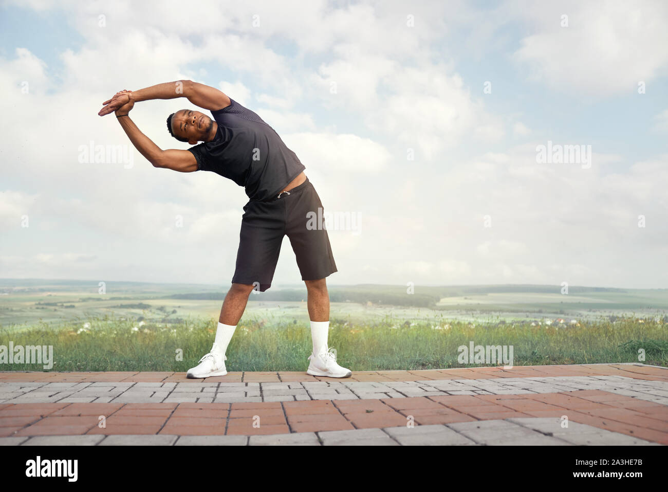 Handsome sportsman making slopes outdoors. Active athlete with muscular body in black t shirt and black shorts doing exercises. Athletic man raising hands, posing on background of blue sky. Stock Photo