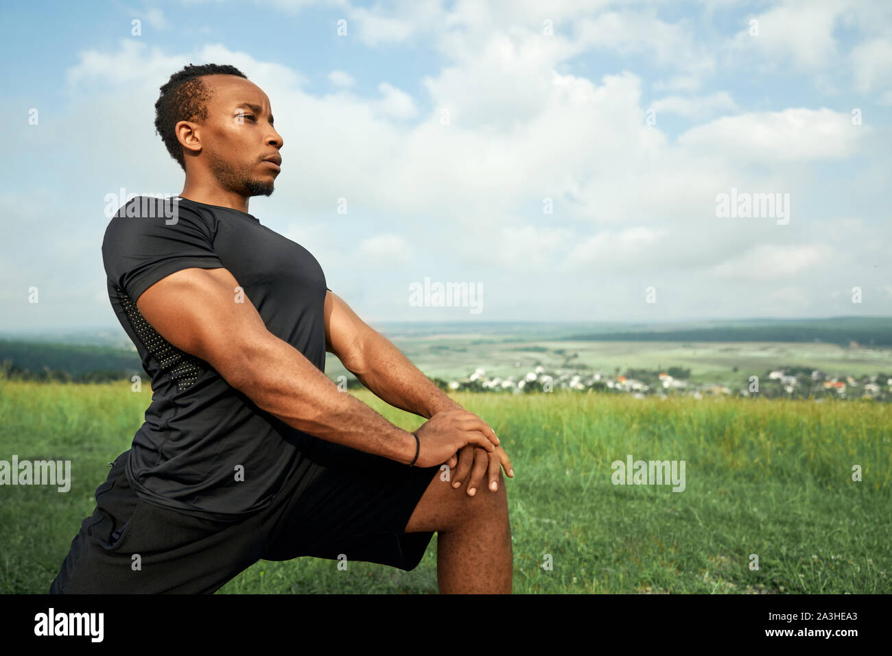 Handsome african man doing exercises in morning outdoors. Sportsman with muscular body wearing in black t shirt and shorts stretching. Athlete posing on background of blue sky. Stock Photo