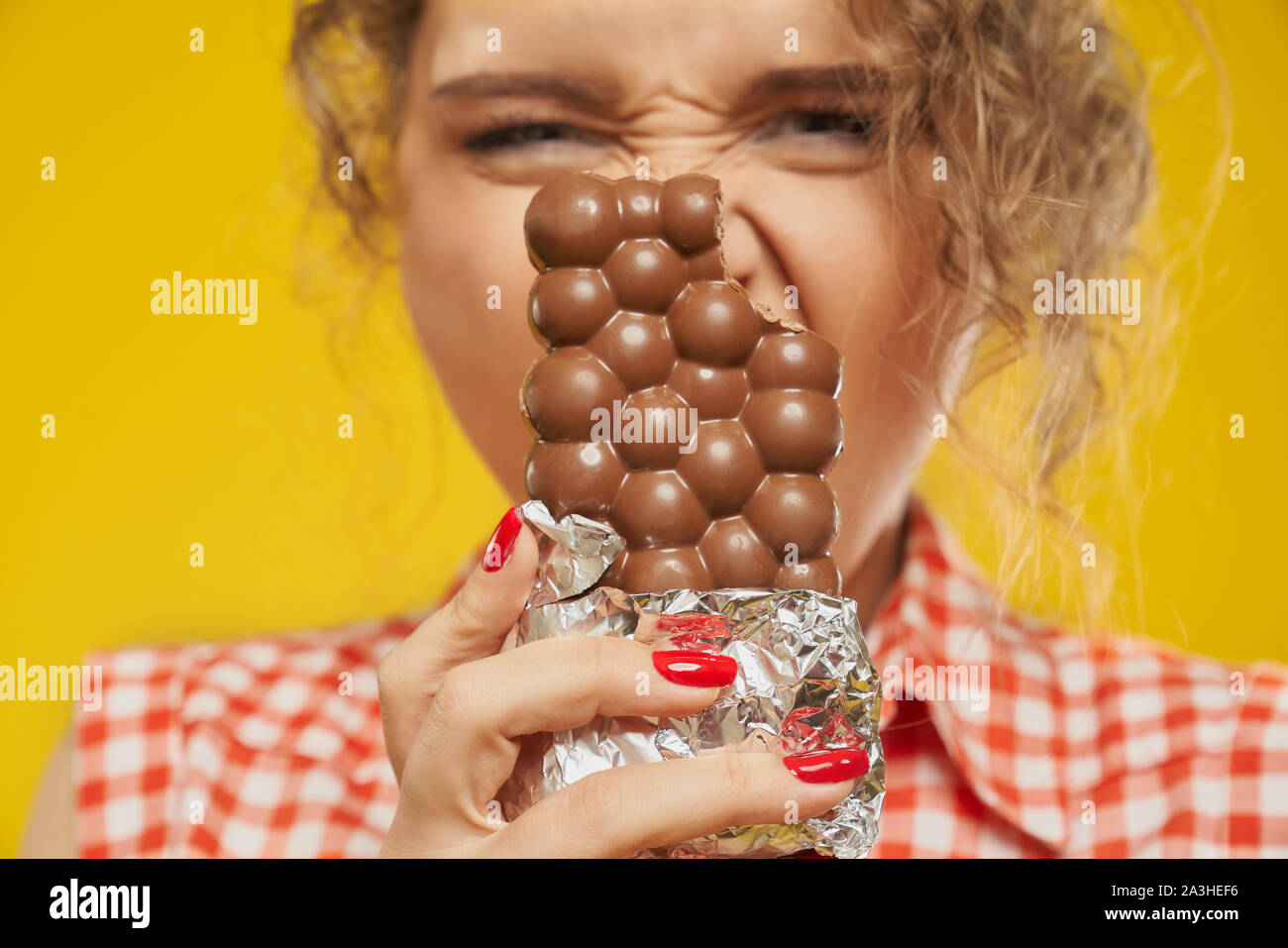 Close up of milk bubbly chocolate bitten from one side that keeping young blonde girl with curly hair and red nails over yellow background. Concept of temptation and pleasure during eating sweets Stock Photo