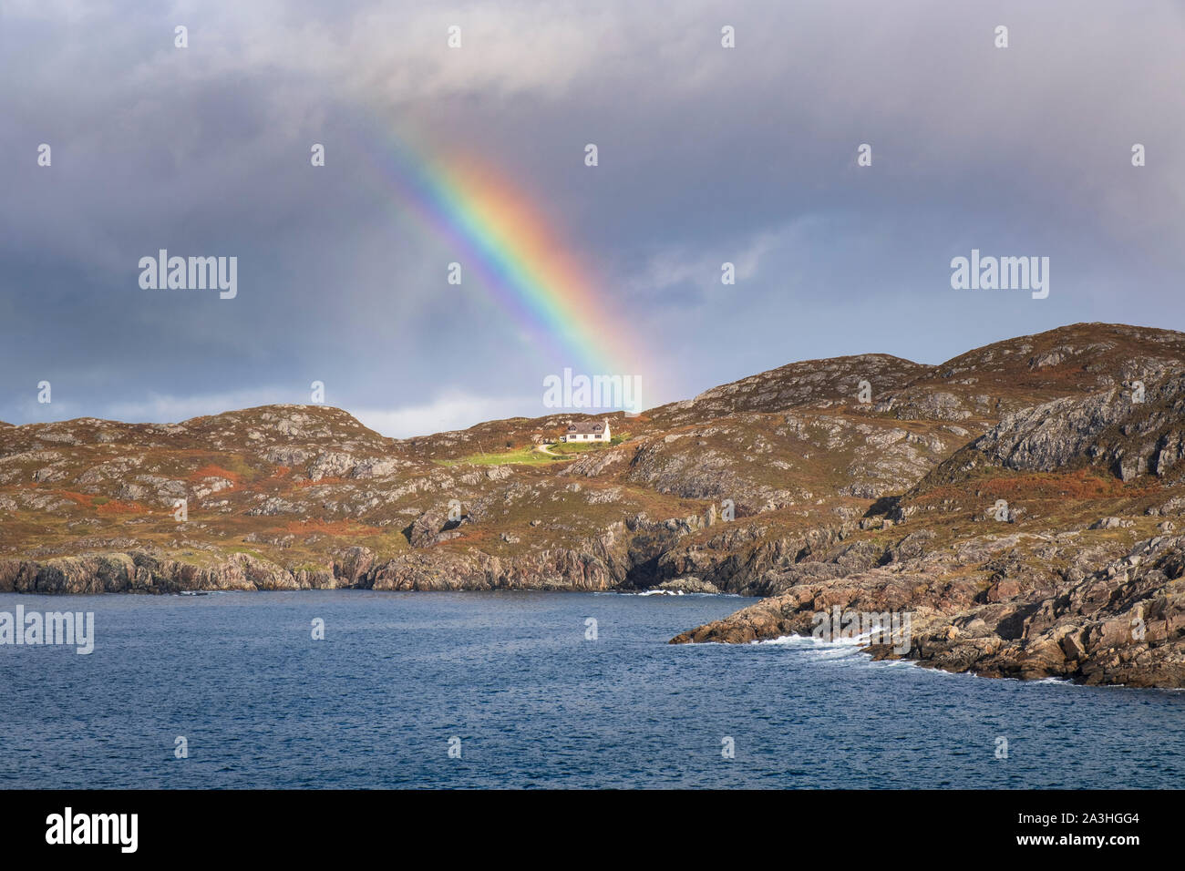 A home on the coast somewhere over the rainbow a lone cottage or house by the sea on a remote barren coastline with dark cloudy sky and bright rainbow Stock Photo