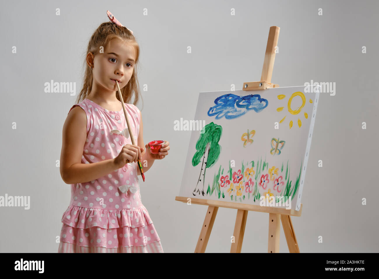 Little girl artist in a pink dress is standing behind easel and ...