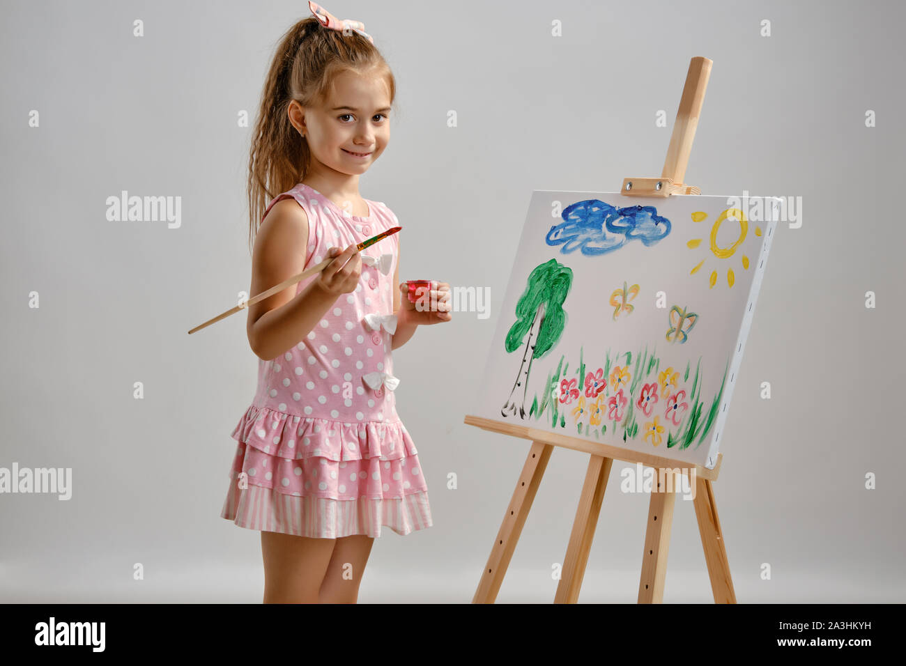 Little girl artist in a pink dress is standing behind easel and ...