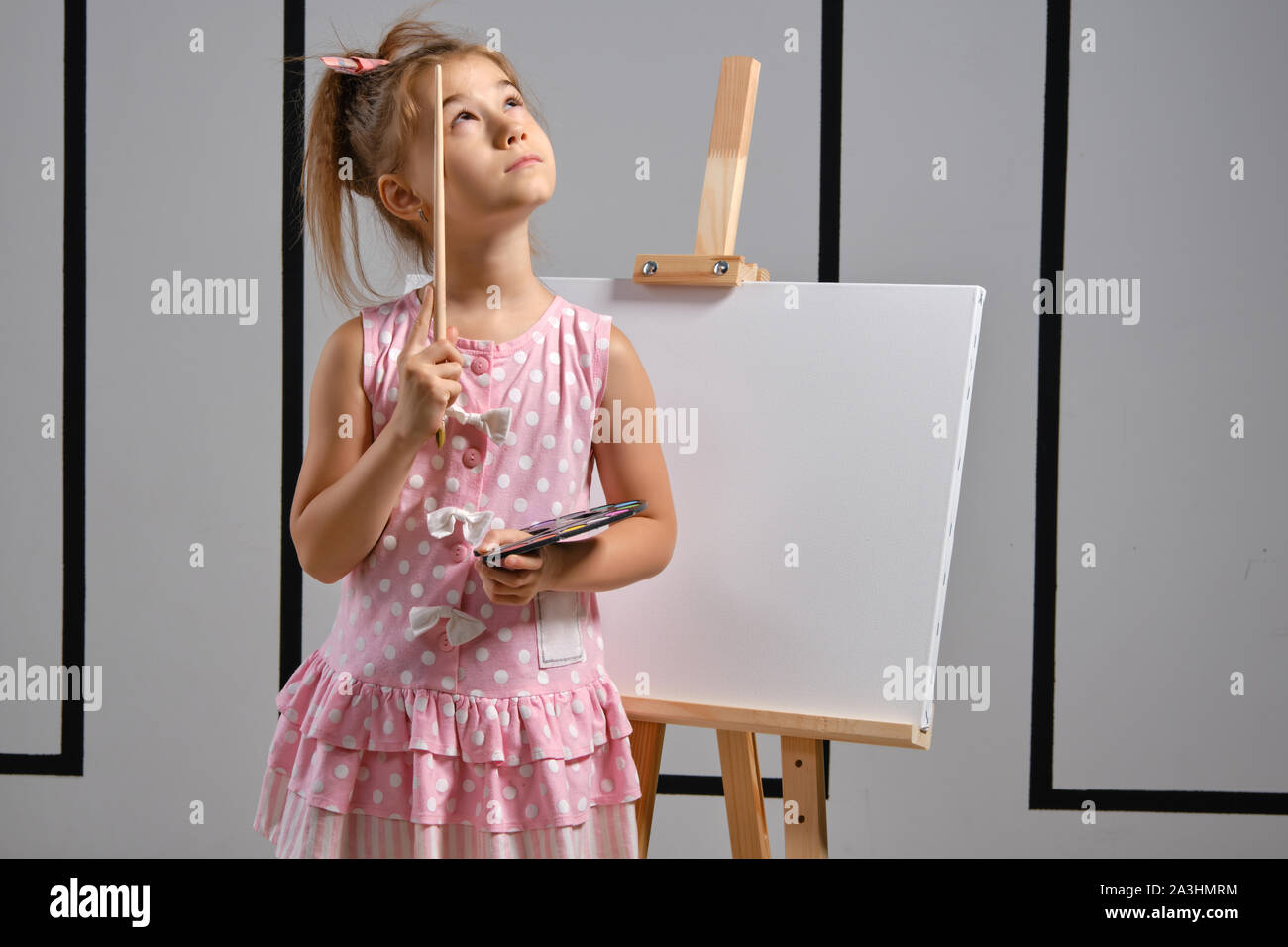 Little girl artist in a pink dress is standing behind easel and ...