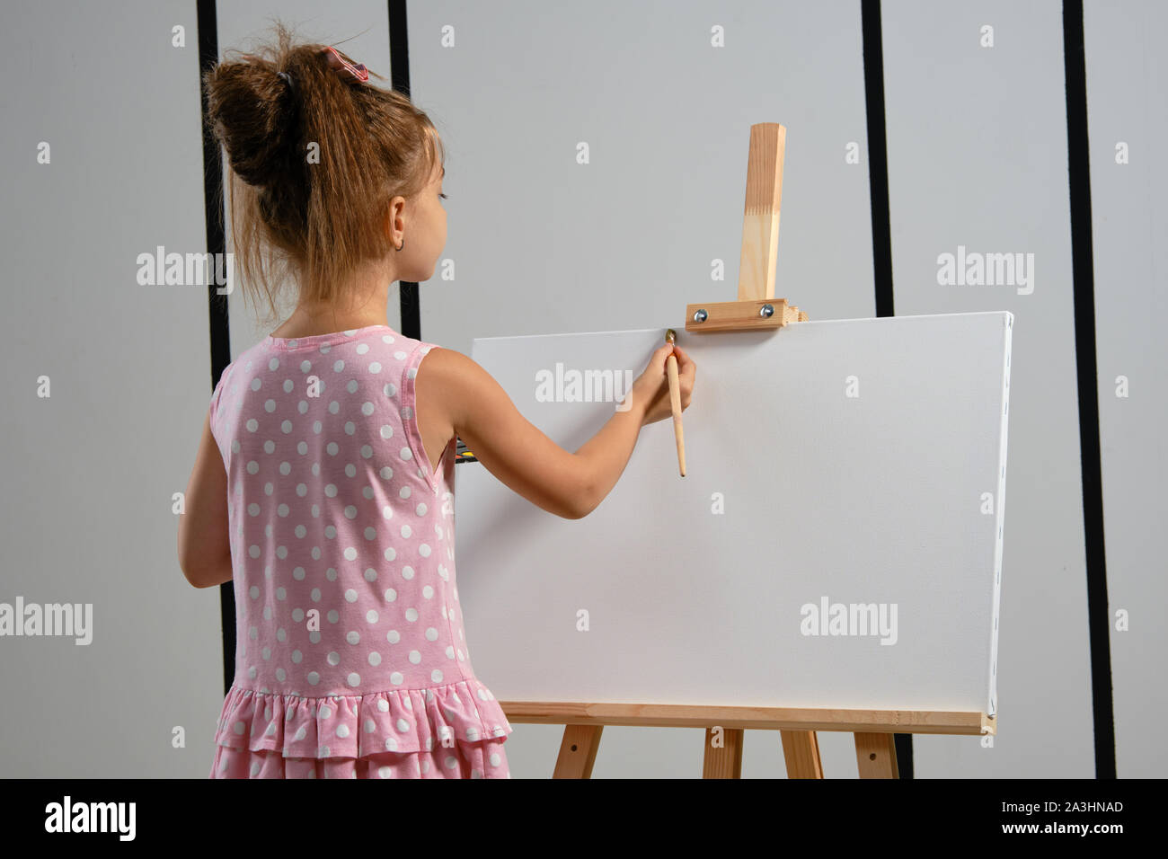 Little girl artist in a pink dress is standing behind easel and ...