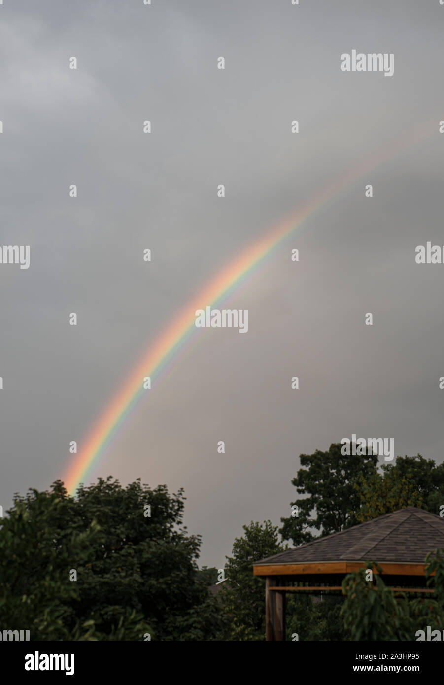 Bright rainbow over some trees in a dark cloudy sky. Stock Photo
