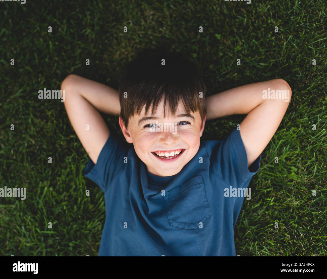 Overhead close up of happy boy laying on grass with arms behind head. Stock Photo