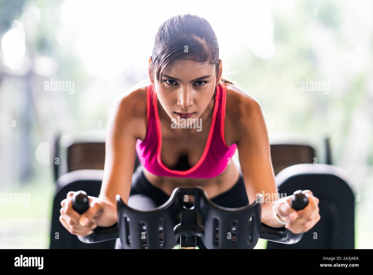 Beautiful Asian woman exercising on stationary cycling machine in indoor fitness gym, determination face. Sport recreational activity, people workout Stock Photo