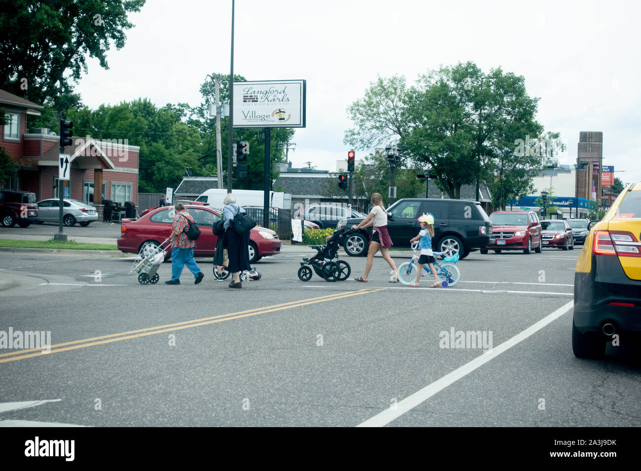 Children and adults with grocery cart bike tricycle pram crossing a busy street intersection with traffic lights. St Paul Minnesota MN USA Stock Photo
