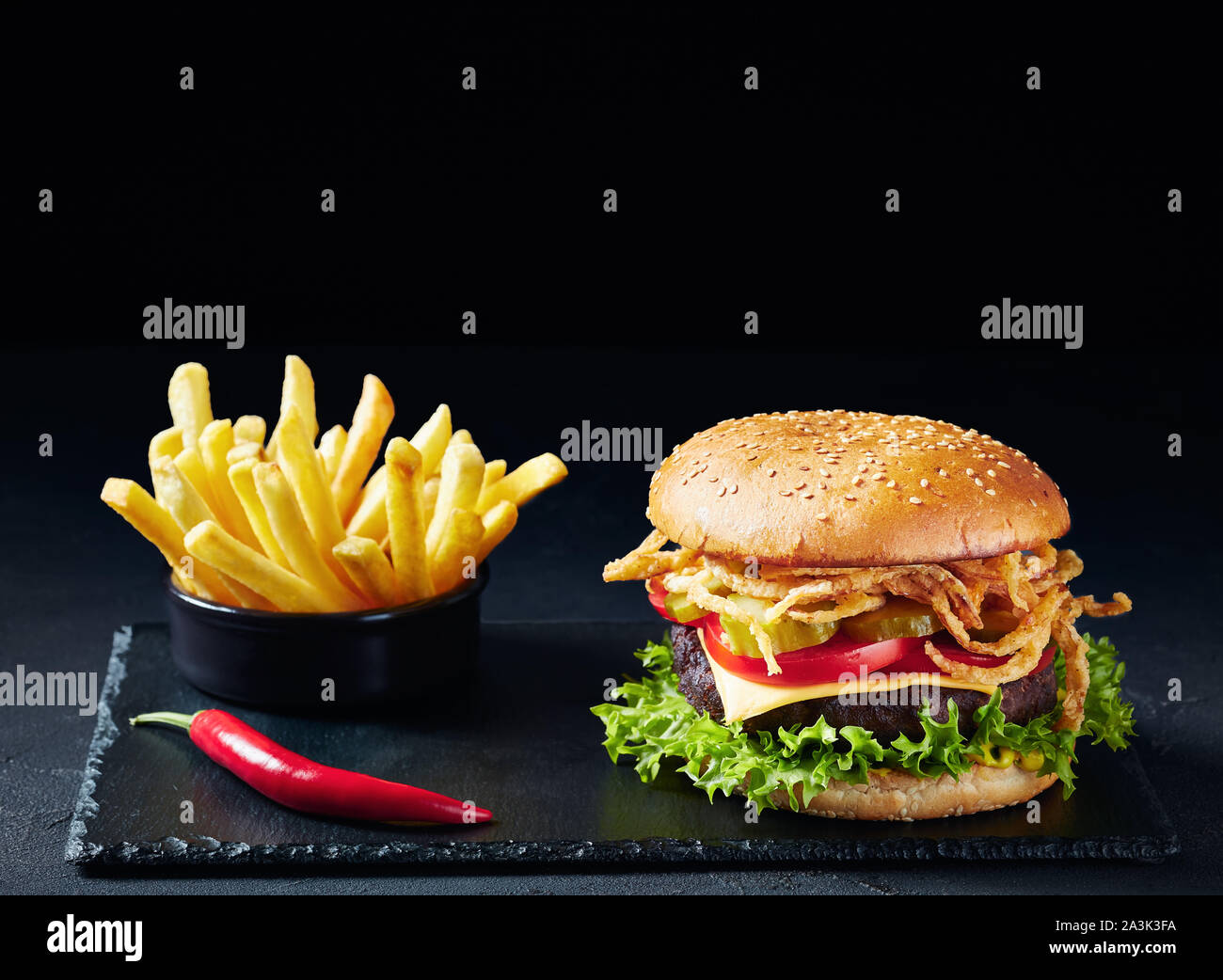 Cheeseburger with Beef Patty, cheddar cheese, crispy fried onions, lettuce, sliced tomatoes pickled cucumbers on a black stone board with french fries Stock Photo