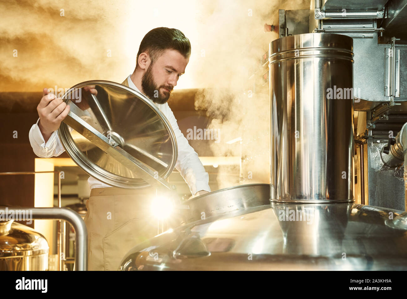 Brewery specialist opening cover of brew kettle, looking down. Engineer inspecting process of brewing beer with steam. Concept of modern bavaria. Stock Photo