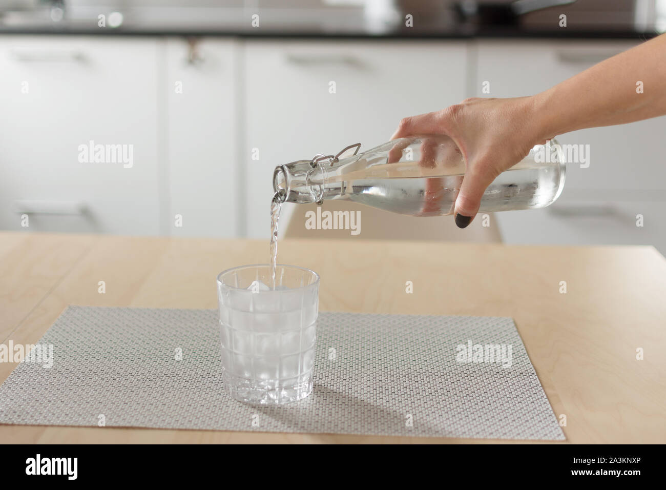 Female hand pouring cold water from bottle into glass with ice cubes on wooden table over kitchen background. Stock Photo