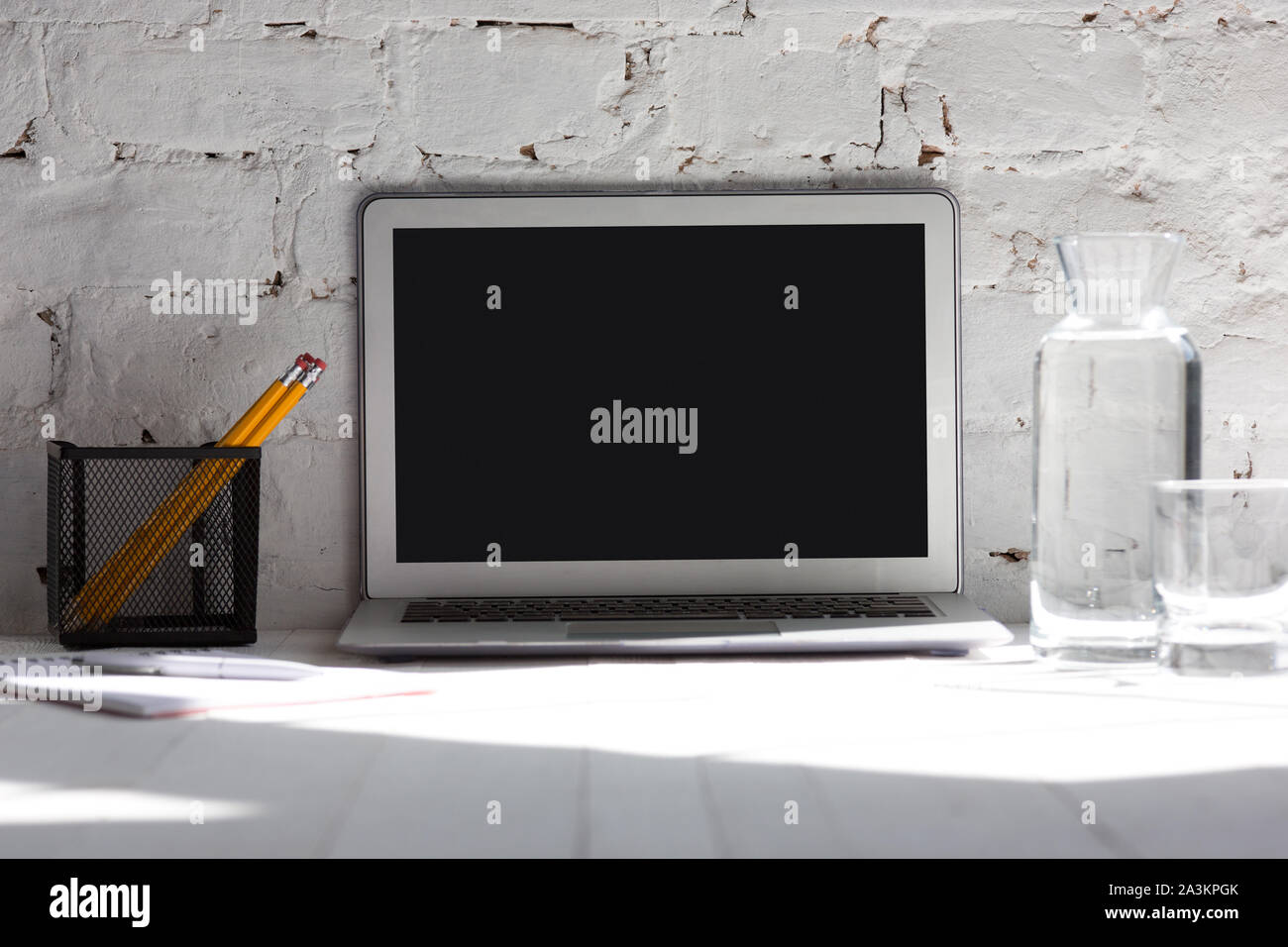 Mock up empty black laptop screen on white brick wall background with pencils, notes and glass of water. Copyspace, negative space for your advertisin Stock Photo