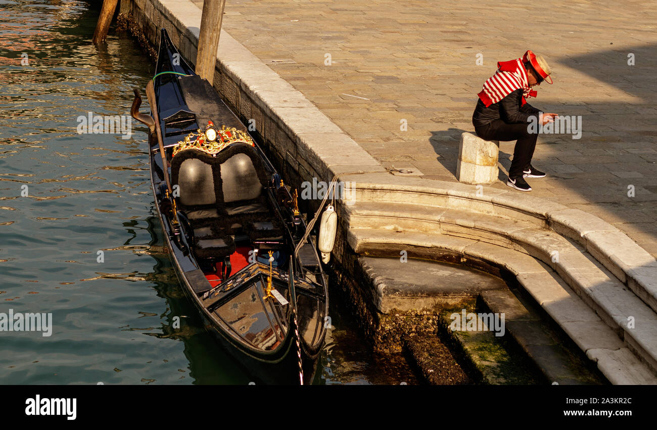 Venice / Italy - February 16 2018: Gondolier sitting next to a Gondola studying his mobile phone while wating for tourists Stock Photo