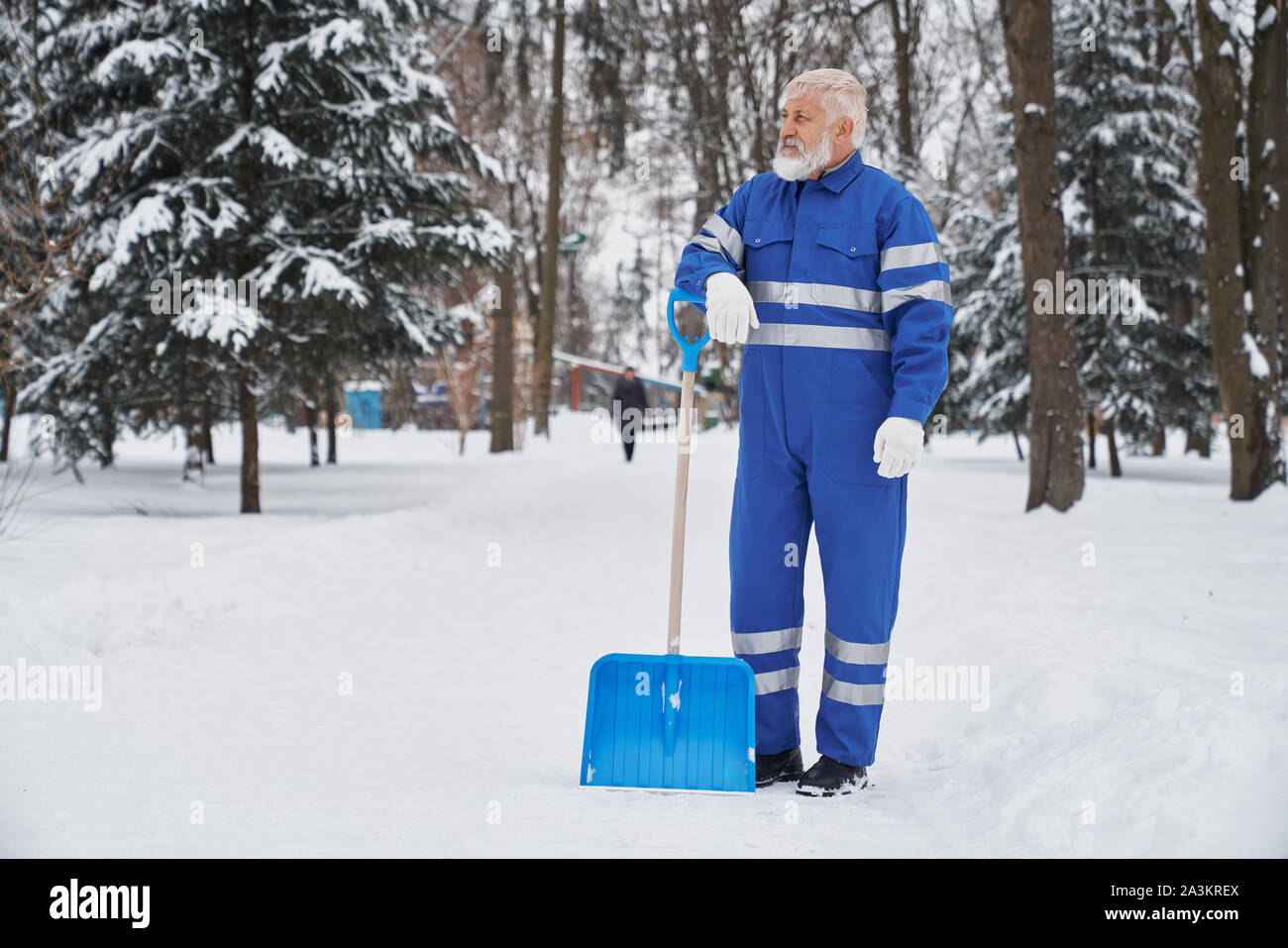 Male janitor in blue uniform standing in park and resting while cleaning sidewalks from snow. Male worker keeping equipment and looking aside in winter. Concept of clearing and service. Stock Photo