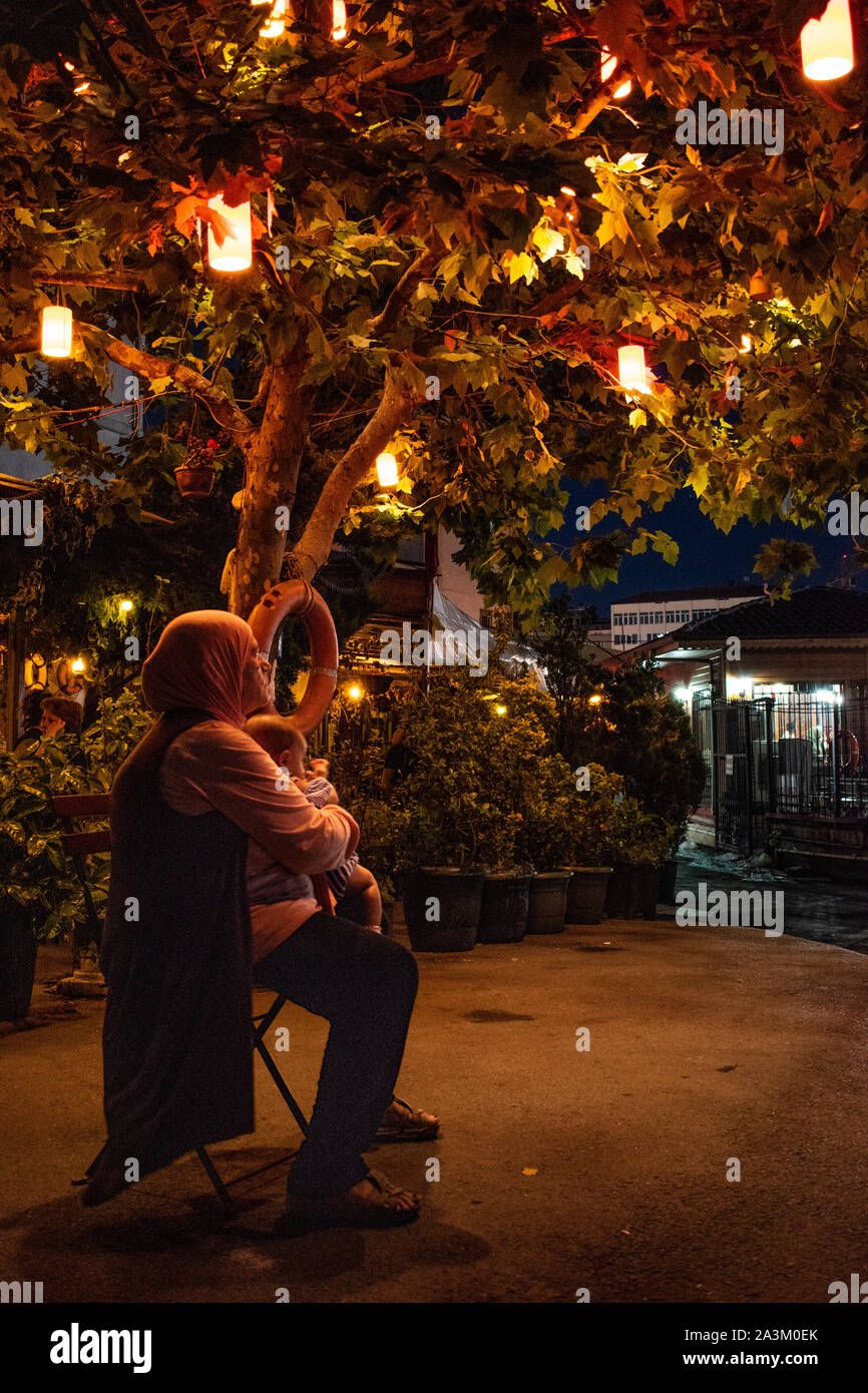 Istanbul: nightlife in the city, a Turkish woman seated on a chair under a tree decorated with colored lanterns and cradling a baby in the streets Stock Photo