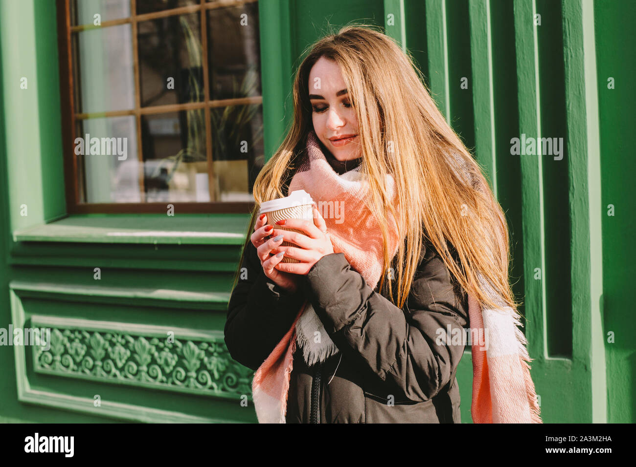 Happy woman with disposable cup in warm clothing standing beside green building in city street during winter Stock Photo