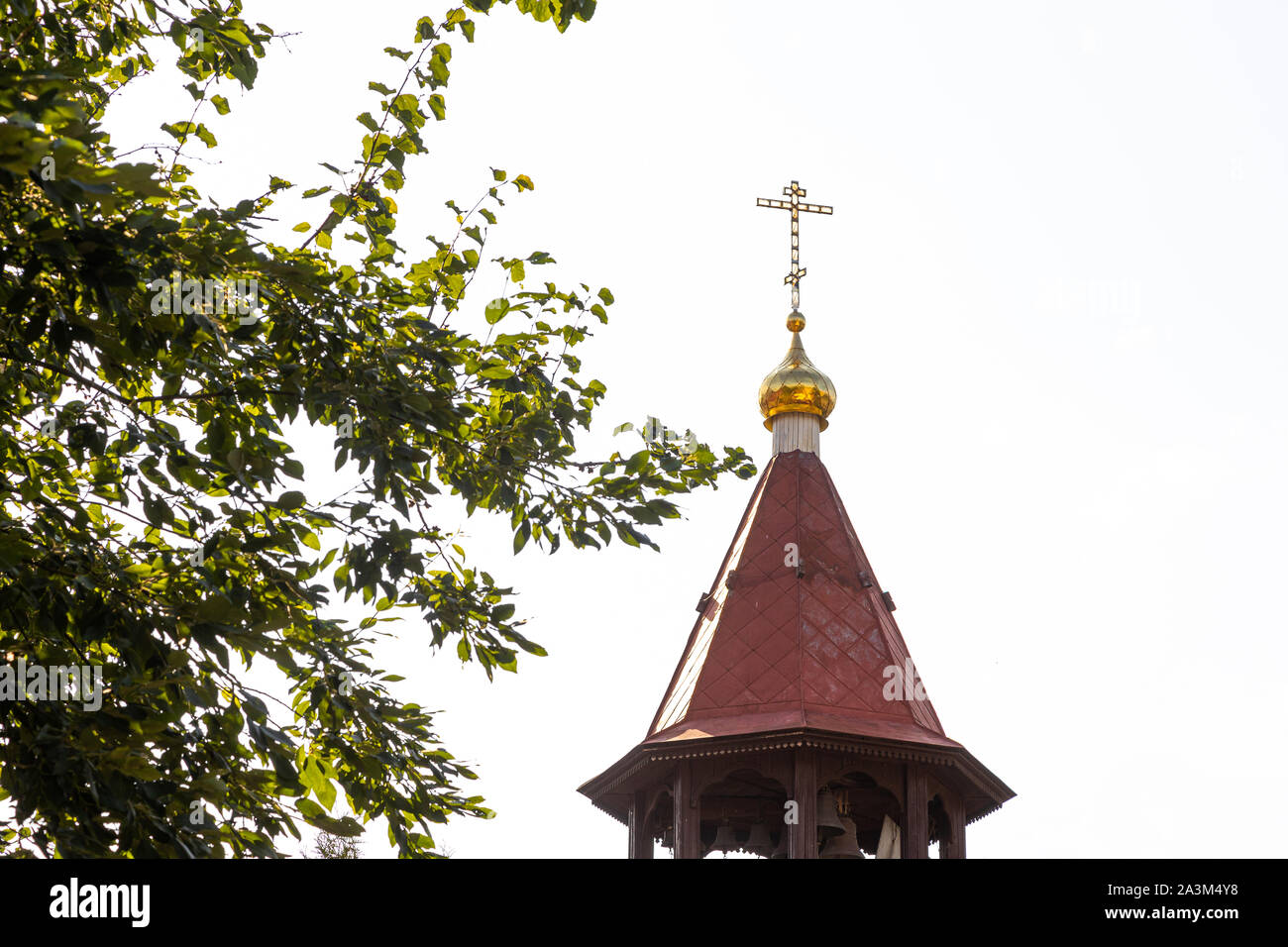 Maroon top of orthodox church bell tower with cross against sky and tree leaves Stock Photo