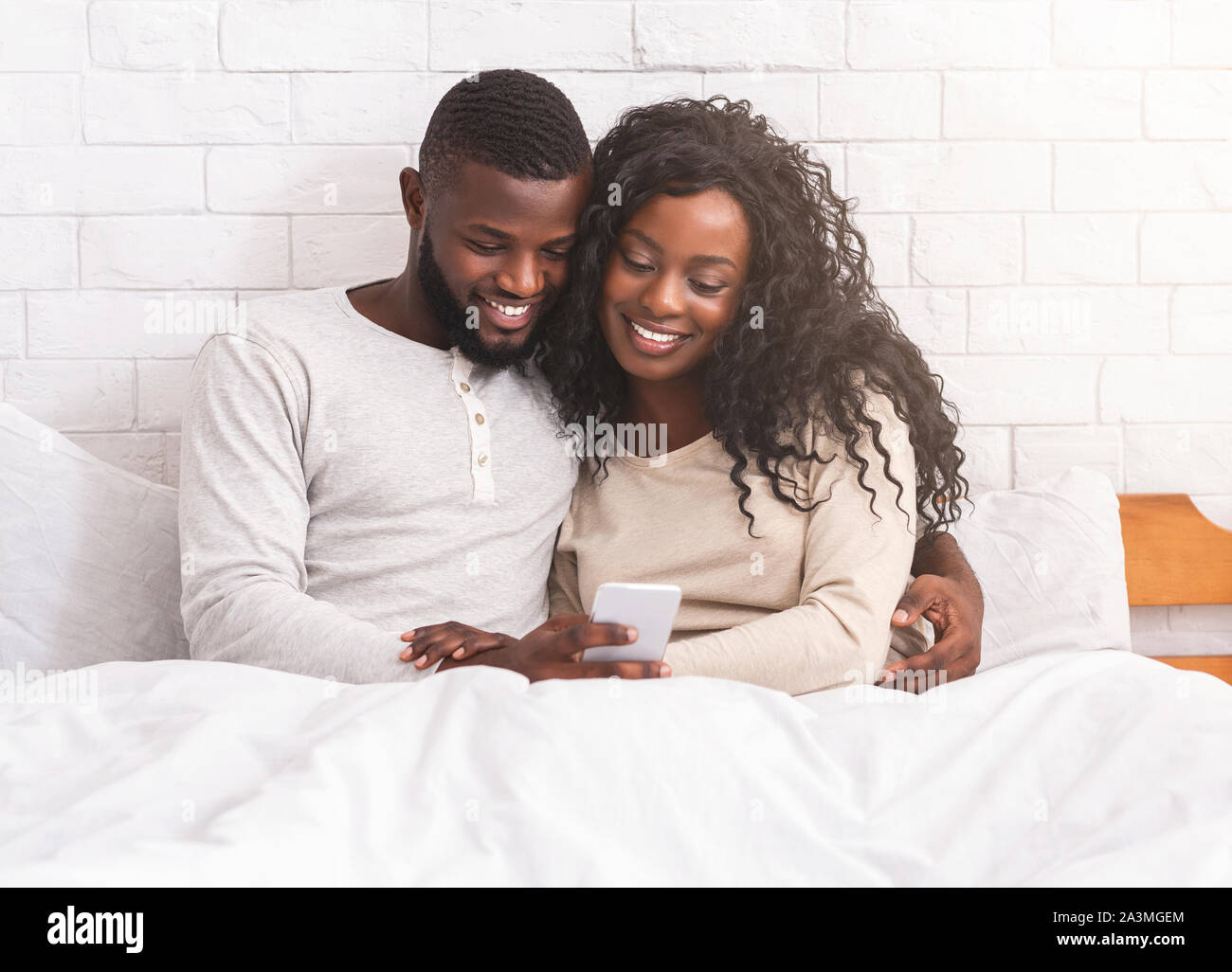 Smiling black couple using smartphone sitting on bed together Stock Photo