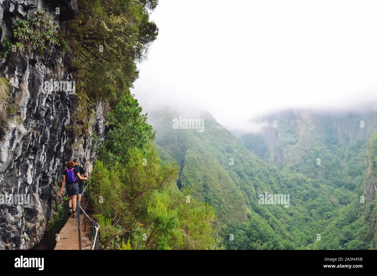 Hikers walking on a narrow path on the edge of the rock during Levada do Caldeirao Verde Trail. Misty green mountains in background. Dangerous hiking. Portuguese tourist attraction. Fog, foggy. Stock Photo
