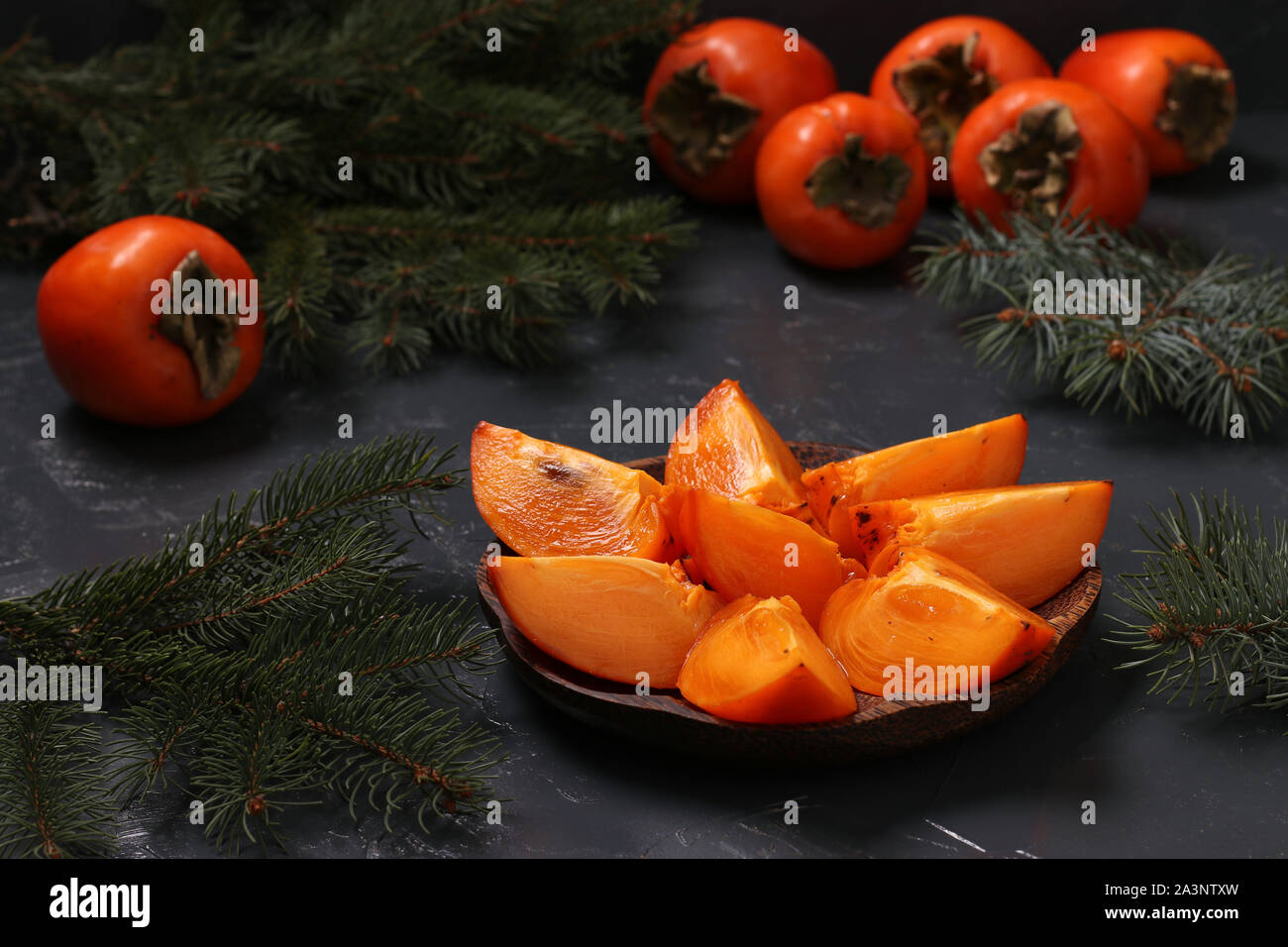 Slices of ripe orange persimmon are on a wooden plate on a dark background, horizontal orientation, closeup Stock Photo