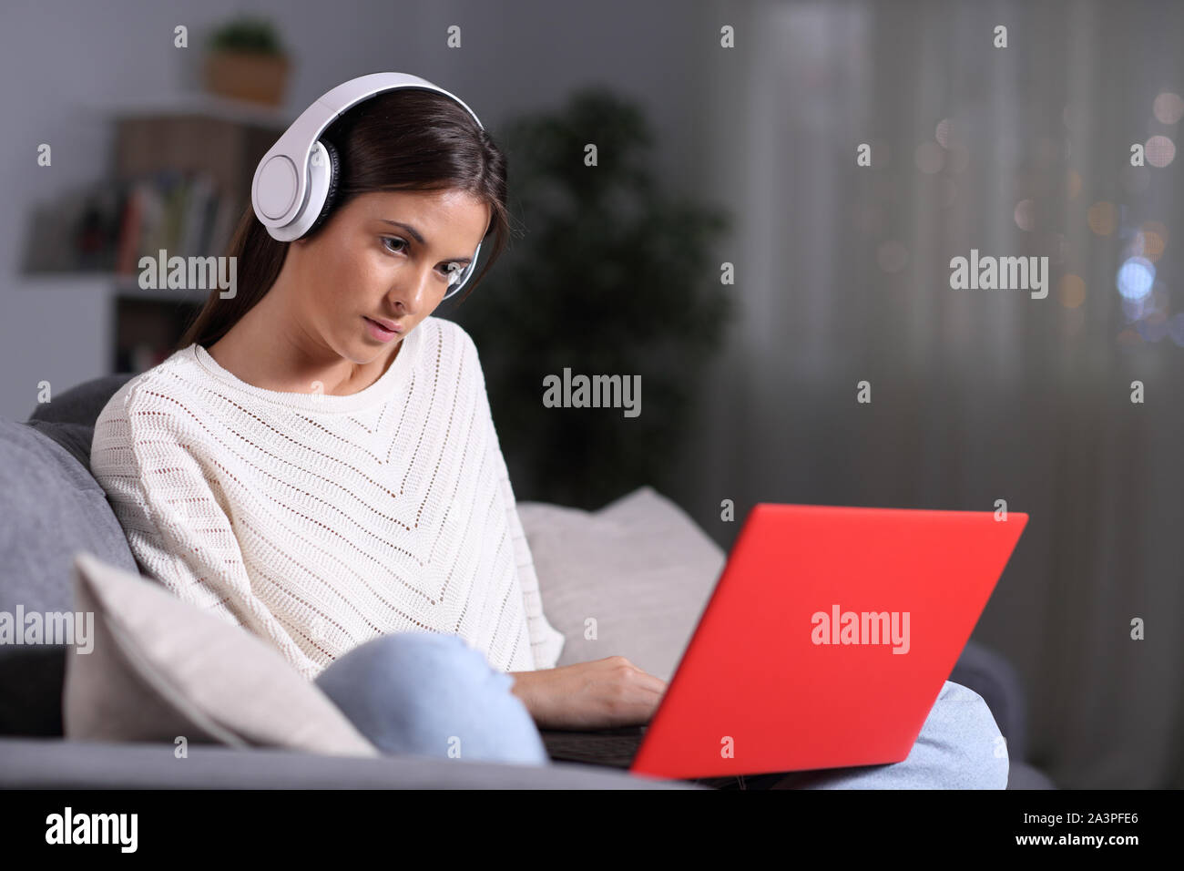 Serious girl wearing headphones checking red laptop content sitting on a couch in the night at home Stock Photo