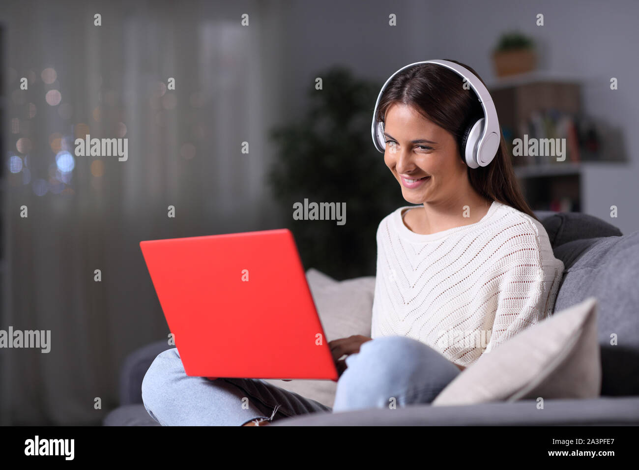 Happy girl wearing headphones using a red laptop sitting on a couch in the night at home Stock Photo