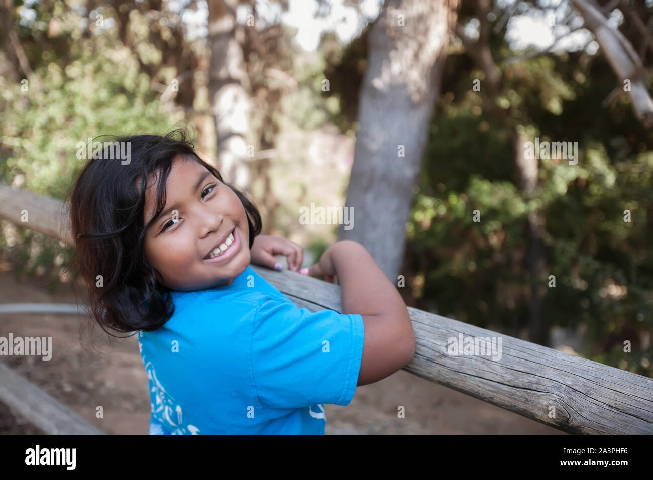 A little girl with a big smile who is holding on to a wooden fence and surrounded by trees in a natural environment. Stock Photo